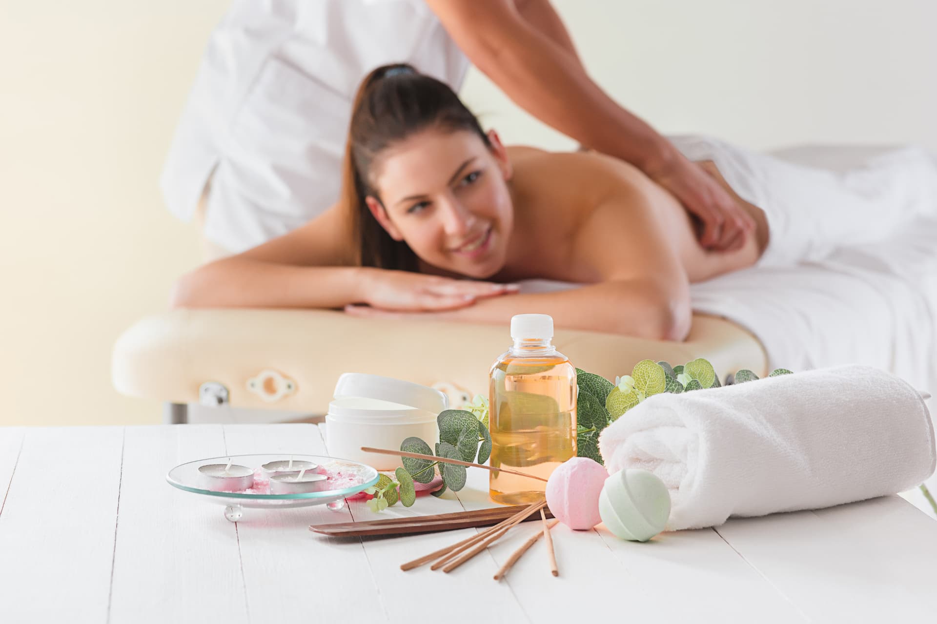 A happy young woman lies on a massage table and smiles. In the foreground are massage utensils.