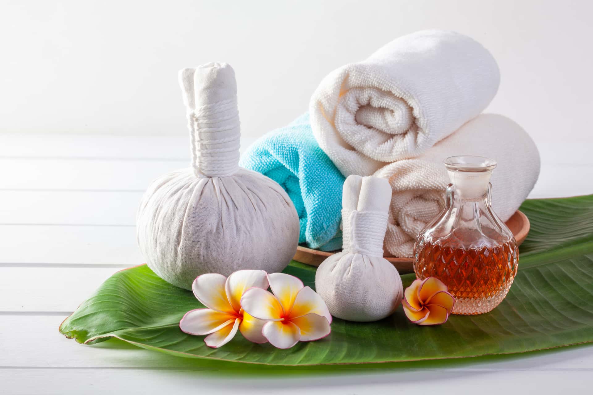 Small selection of herbal stamps, massage oil and towels arranged on banana leaf.